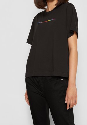 Woman wearing a black t-shirt with a small multicolored wavy line design on the chest, paired with black pants.