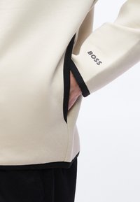 Μπεζ μακρυμάνικο ένδυμα με μαύρο περιθώριο στα μανίκια και στο τελείωμα, χέρι στην πλαϊνή τσέπη, λογότυπο "BOSS" στο μανίκι κοντά στον καρπό.