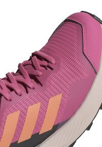 Zapatilla deportiva rosa con material texturizado, con franjas naranjas, diseño con cordones y una suela de goma duradera.