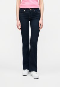 Γυναίκα με σκούρα μπλε τζιν παντελόνι καμπάνα, λευκά αθλητικά παπούτσια και ροζ κοντομάνικο πουκάμισο, στέκεται μπροστά σε απλό φωτεινό φόντο.