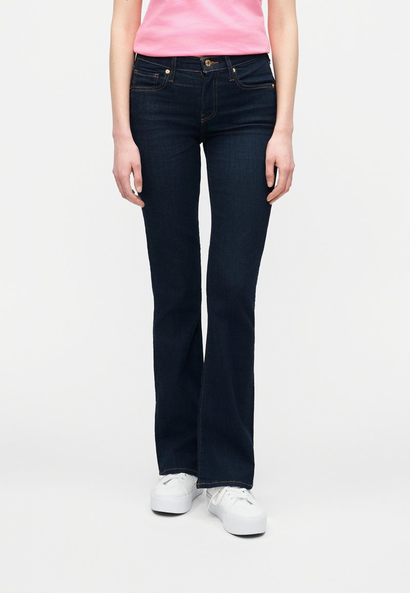 Γυναίκα με σκούρα μπλε τζιν παντελόνι καμπάνα, λευκά αθλητικά παπούτσια και ροζ κοντομάνικο πουκάμισο, στέκεται μπροστά σε απλό φωτεινό φόντο.