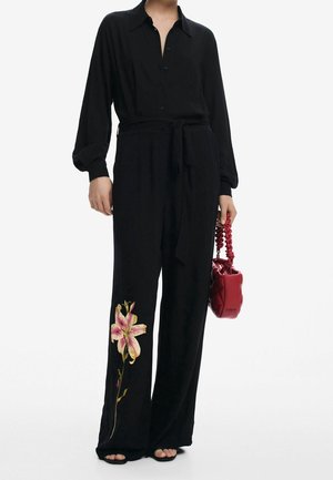 Mono de seda negra de manga larga con un frente abotonado, cintura ceñida con cinturón, pantalones de pierna ancha y bordado floral en rosa y verde en la pierna izquierda. Lleva un bolso rojo.