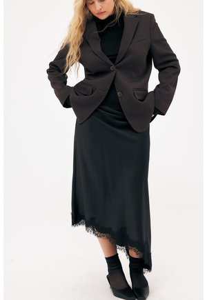 Kobieta ubrana w czarną marynarkę na czarnym golfie i czarną asymetryczną spódnicę z koronkowym wykończeniem, do tego czarne skarpetki i szpiczaste buty.