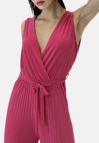 Mono sin mangas de color rosa con un escote en V profundo, textura tejida, diseño plizado y un cinturón atado en la cintura. Tela suave y fluida.