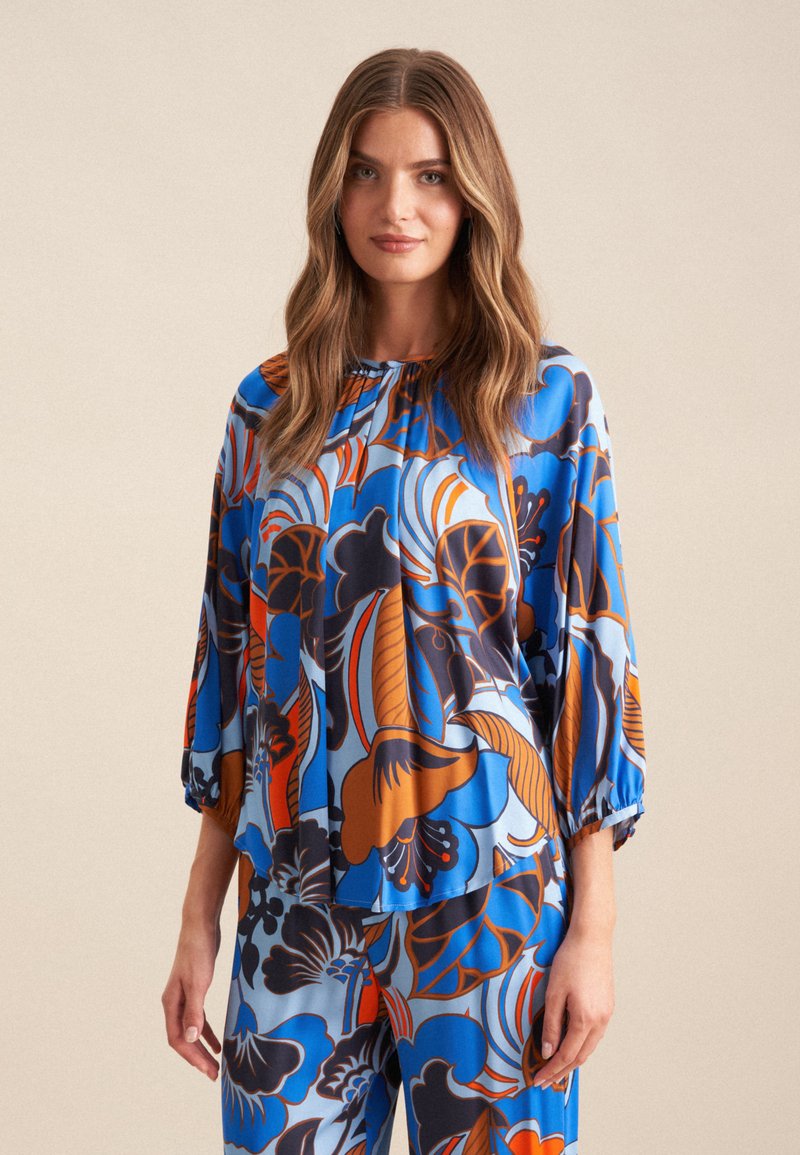 Blusa floreale blu con maniche a palloncino, caratterizzata da motivi arancioni e scuri. Tessuto morbido e fluido. Indossata con pantaloni coordinati.