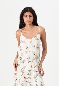 Rochie cu imprimeu floral dintr-un material crem deschis, având bretele subțiri tip spaghetti, decolteu în V și talie ajustată cu șnururi.