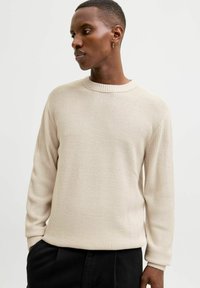 Pull en tricot beige avec un motif texturé, col rond et manches longues, accompagné d'un pantalon noir. Manches avec des poignets doux en côtes.