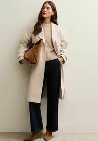 Abrigo trench beige sobre un suéter claro, combinado con jeans oscuros de pierna ancha y tacones marrones. Un bolso marrón de ante completa el look.