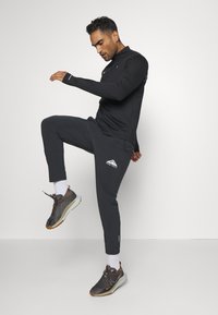 Schwarzes langärmliges Sportoberteil mit hohem Kragen, kombiniert mit schwarzen taillierten Hosen mit einem Logo. Dunkle Sneakers mit orangefarbenen Akzenten runden das Outfit ab.