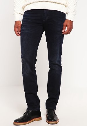 Homme portant un jean slim bleu foncé, des bottes en cuir noires et un pull en maille blanc, debout devant un fond uni.