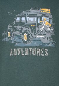 Donkergroene t-shirt met een grafisch ontwerp van een zwarte off-road voertuig dat een motorfiets vervoert, met daaronder de tekst "AVONTUREN" in vetgedrukte, gestreepte letters.