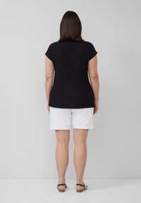 Zwarte, gestructureerde top met korte mouwen, gecombineerd met witte shorts. Sandalen met een bruine houten zool zichtbaar. Effen, lichtgrijze achtergrond.