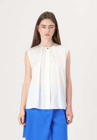 Mouwloze witte blouse van lichtgewicht stof, met een geplooide voorkant en een kleine knop bij de hals.
