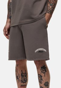 Shorts in cotone grigio con una vestibilità rilassata, caratterizzati da un logo bianco sulla coscia sinistra e un orlo cucito. La texture è morbida e leggermente spazzolata.