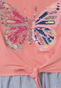 Różowy materiał z dużym wzorem motyla wykonanego z różowych, srebrnych, niebieskich i żółtych cekinów, związany w węzeł nad materiałem w niebiesko-białe paski.
