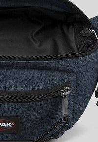 Blå tygpås med en текстурированная yta, svart dragkedjestängning och en liten logolapp. Inuti är den svart med tygfoder.