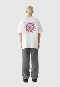 Bela oversize majica s abstraktnim grafičkim vzorcem in besedilom na hrbtni strani. Nosijo jo z sivimi jeans hlačami in črnimi športnimi copati.