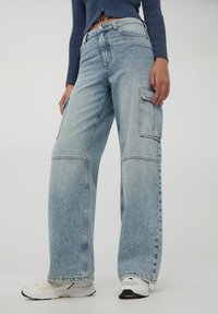 Jasnoniebieskie dżinsowe spodnie cargo z szerokimi nogawkami, przednimi i bocznymi kieszeniami oraz kontrastowymi szwami. Noszone z białymi butami sportowymi.