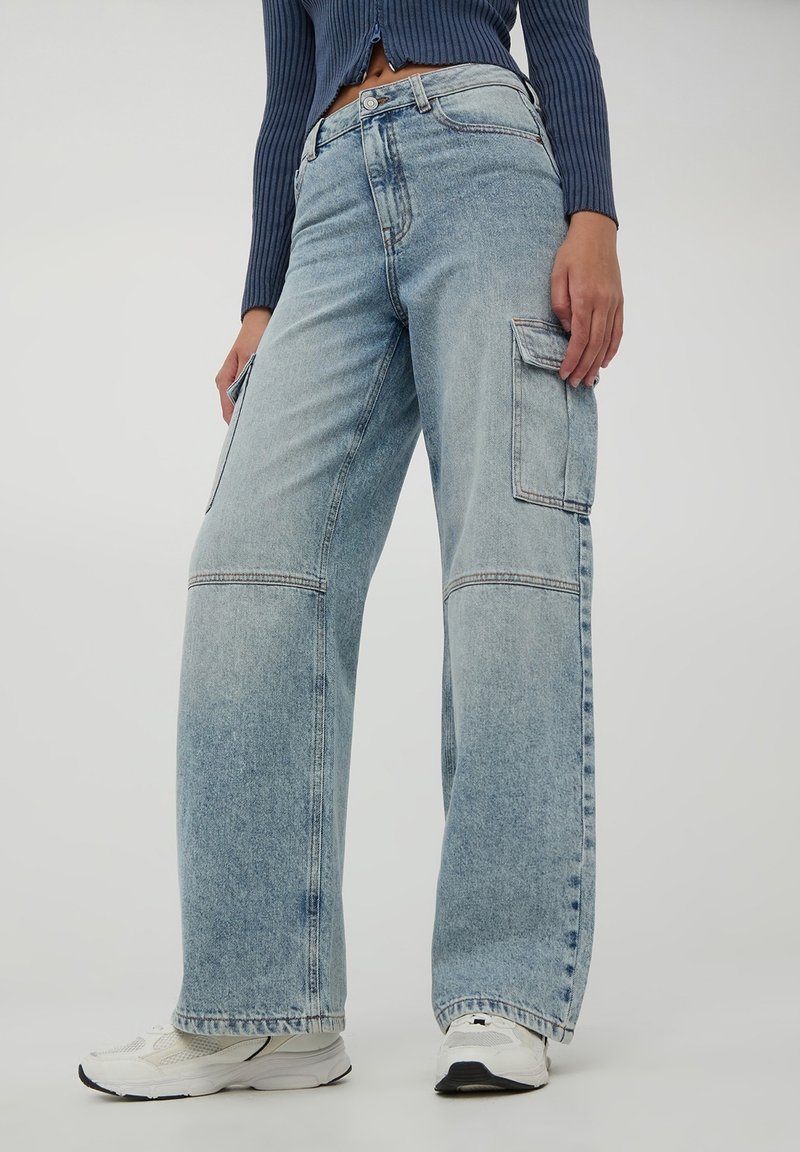 Jasnoniebieskie dżinsowe spodnie cargo z szerokimi nogawkami, przednimi i bocznymi kieszeniami oraz kontrastowymi szwami. Noszone z białymi butami sportowymi.