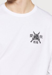 Weißes T-Shirt mit einem schwarzen Emblem, das zwei gekreuzte Schwerter, ein wirbelndes Schild und vier asiatische Zeichen in der Nähe des linken Brustbereichs zeigt.