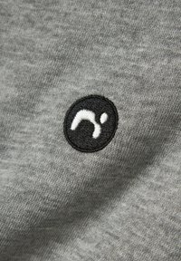 Logo ricamato in bianco e nero con una forma curvata e un punto su uno sfondo di tessuto grigio, vista ravvicinata.