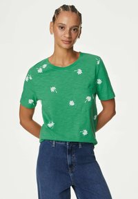 Camiseta de algodón verde con mangas cortas, con bordado floral blanco y un pequeño bolsillo en el frente. Cuello redondo sencillo y corte holgado.