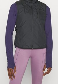 Svart lättviktsväst med hög krage, dragkedjestängning och en liten bröstficka. Bärs över en lila långärmad tröja och rosa leggings.