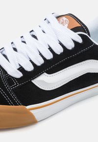 Tenisi negri din suede cu sireturi albe și dungi pe lateral, talpă din cauciuc de tip gumă și detalii de cusătură albă. Prezintă un tab cu logo portocalie pe toc.
