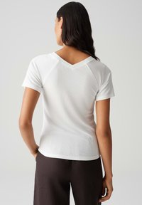 Weißes, geripptes T-Shirt mit kurzen Ärmeln, V-Ausschnitt und tailliertem Schnitt. Die Rückseite hat eine subtile Nahtdetails, kombiniert mit dunklen Hosen.