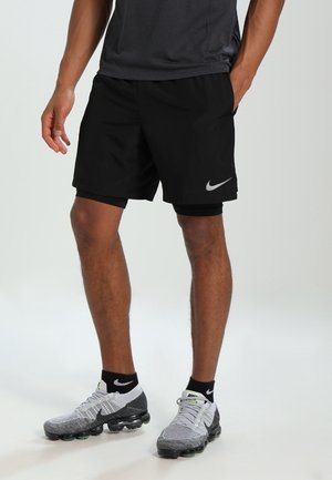 Schwarze Sportschuhe aus leichtem Stoff, mit elastischem Bund und einem silbernen Nike-Logo. Kombiniert mit grauen Nike-Sneakern.