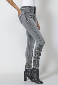 Šedé džíny z denimu s detailním černým květinovým vyšíváním na lemech, slim fit, vysoký pas a třepením na okrajích. V kombinaci s černými botami na podpatku.