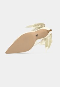 Chaussure à talon avec une tige crème, un bout pointu et de douces accents en satin. Elle dispose d'une semelle beige et d'un nœud décoratif avec un détail en métal.