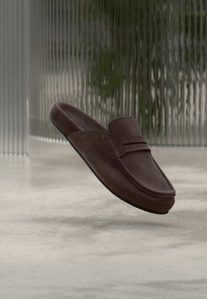 Mocasín marrón de ante sin cordones flotando sobre un suelo gris liso con un panel de vidrio texturizado y vegetación al fondo.