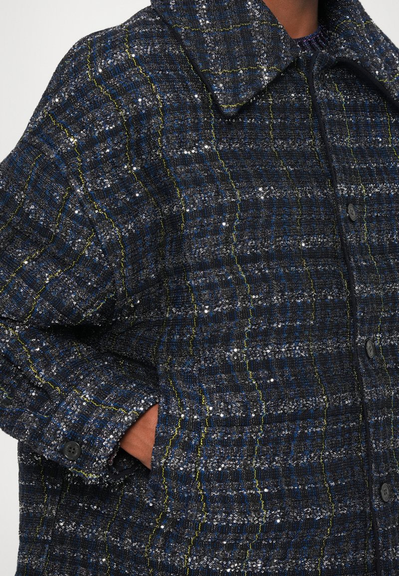 Navyblauer gewebter Mantel aus strukturiertem Stoff mit silbernen Pailletten und grünen Akzenten, großem Kragen und Knöpfen. Enthält Seitentaschen.