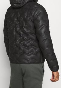 Sort pufferjakke med quiltet design og shiny overflade. Har en hætte og ribbet kant, parret med grønne sweatpants.