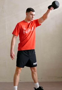 Punane lühikeste varrukatega sportlik särk valge logoga, mustad šortsid valgete kiri, tõstmas musta kettlebelli heleda taustaga.