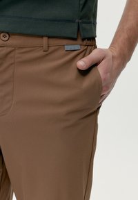 Tuvplānā redze uz personas roku brūnās bikses priekšējā kabatā ar pogu un mazu birku, kas nēsā tumši zaļu kreklu.