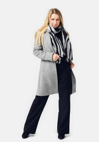 Blonde woman wearing a gray coat, black pants, black top, and a black-and-white striped scarf standing with legs apart.