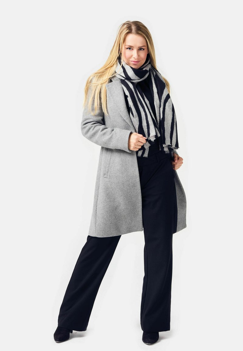 Blonde woman wearing a gray coat, black pants, black top, and a black-and-white striped scarf standing with legs apart.