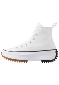 Białe sneakersy za kostkę z płóciennym wierzchem, czarnymi i brązowymi akcentami z gumy, charakterystyczną ząbkowaną solą oraz srebrnymi oczkami z sznurówkami.