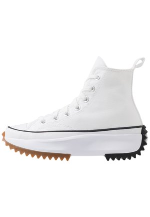 Zapatilla alta blanca con parte superior de lona, acentos de goma negra y marrón, suela dentada destacada y ojales plateados con cordones.