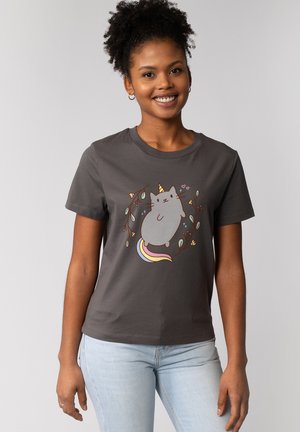 Glunderende vrouw met krullend haar draagt een donkergrijs t-shirt met een grijze katten-unicorn met een regenboogstaart, omringd door bloemrijke elementen.