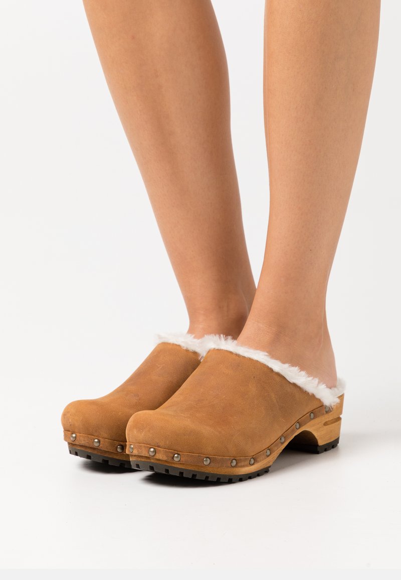 Brown leather clogs with white furry cuff, wooden sole, and black rubber grip on the bottom, featuring decorative metal studs along the edge.