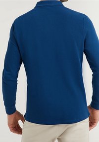 Blå långärmad pikétröja med klassisk krage, mjuk textur och rak fåll. Har en ren, minimalistisk design utan logotyper.