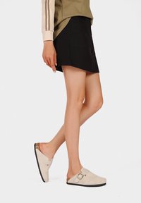 Zoccoli in suede beige con dettaglio a fibbia, upper texturizzato e suola in gomma nera, abbinati a una skort nera e a un top verde oliva.