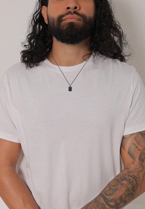 Tricou alb cu mâneci scurte, fabricat dintr-un material moale, purtat cu un colier cu pandantiv negru. Prezintă tatuaje vizibile pe brațe și părul creț.