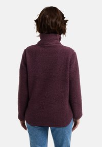Pull en polaire bordeaux avec un col haut, un extérieur texturé, des manches longues et une coupe décontractée, associé à un jean en denim bleu.