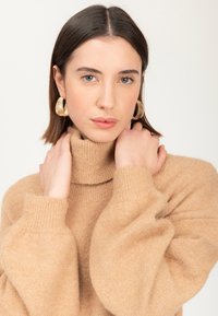 Beige turtleneck sweater med en blød, tekstureret overflade og puffede ærmer; store guldfarvede hoops øreringe tilføjer et metallisk præg.
