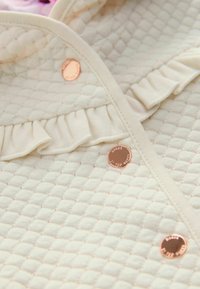 Le tissu matelassé crème clair présente un bord froncé et trois boutons métalliques avec un texte en relief, ajoutant texture et détails.