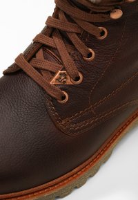 Bota de cuero marrón con superficie texturizada, diseño de cordones, detalles de cobre y suela robusta con un agarre en patrón.
